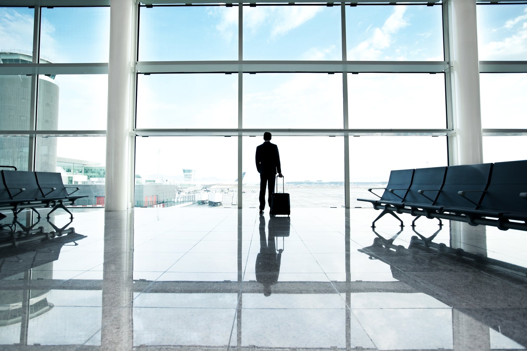 Person in a suit with a carry-on suitcase facing large airport windows