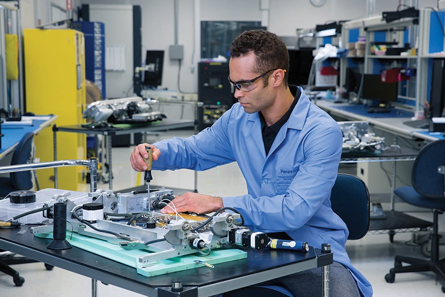 A man in a blue lab coat and safety goggles working at a lab workstation with electronic equipment