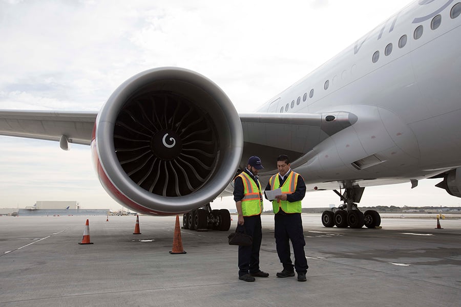 Two airport ground crew members in safety vests stand near a large aircraft engine, discussing a document