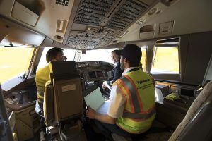 Three men in an airplane cockpit, two seated and one standing, surrounded by control panels and displays